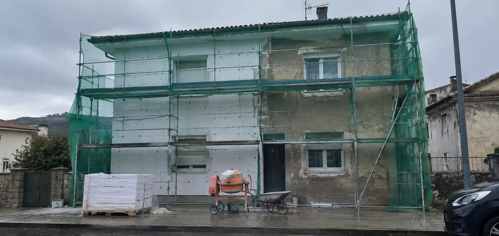 Casa de dos plantas en obras con andamio y malla verde, media fachada ya forrada con placas blancas de aislamiento y la otra mitad con el revoco viejo, con un palé de placas, una hormigonera y una carretilla en la acera mojada
