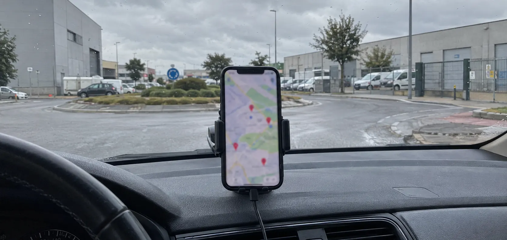 Móvil en el soporte del salpicadero con un mapa y varias chinchetas rojas, en un coche parado ante una rotonda de un polígono industrial en un día de lluvia